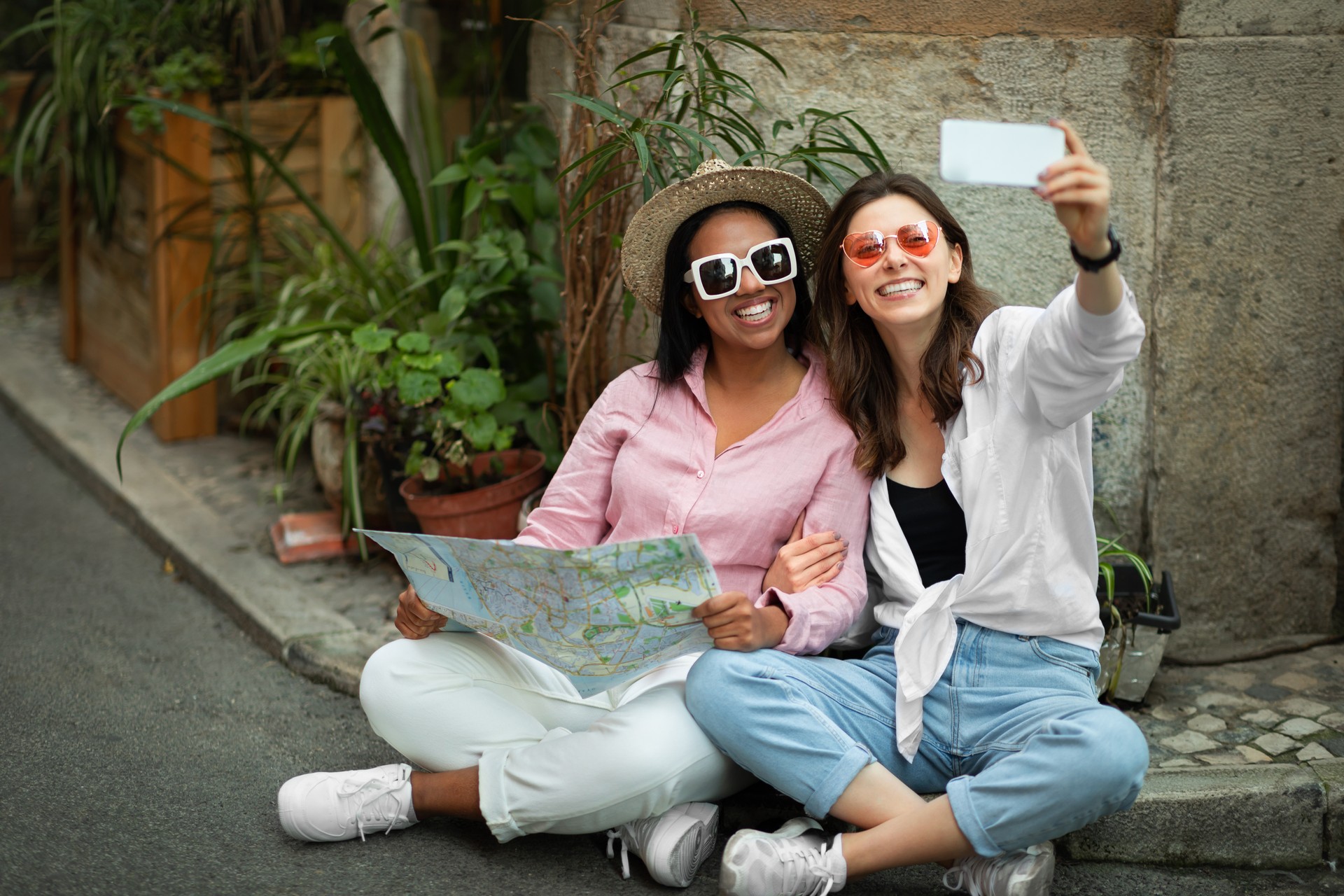 Cheerful millennial multiracial women tourists in casual and hat sitting on floor, looking at map, taking selfie