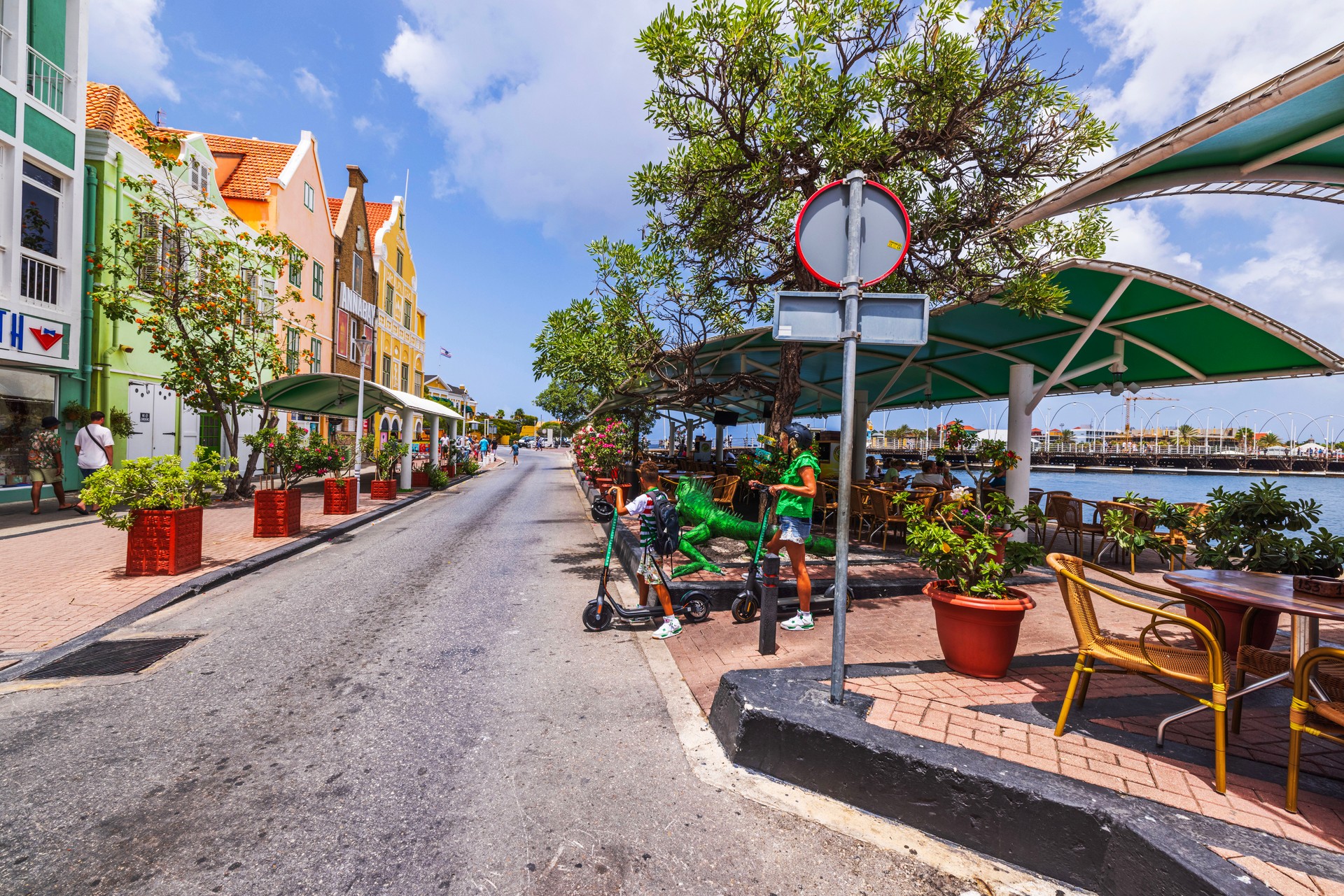 Colorful street view with vibrant buildings, outdoor cafes, and people on scooters near waterfront on sunny day.